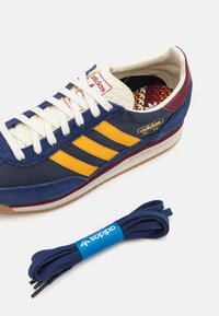 Adidas SL72 kedas ar jūras zila ādas un zamšādas augšdaļu, dzeltenām svītrām, baltām šņorēm un brūnu gumijas zoli. Ietver jūras zila šņores.