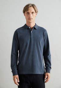Polo cu mânecă lungă, de culoare bleumarin, cu un guler clasic, trei butoane și un broderie simplă a logo-ului pe piept, dintr-un material neted.