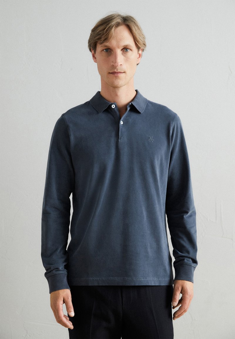 Polo cu mânecă lungă, de culoare bleumarin, cu un guler clasic, trei butoane și un broderie simplă a logo-ului pe piept, dintr-un material neted.