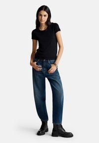 T-shirt nero a maniche corte abbinato a jeans in denim blu a vestibilità rilassata. Il modello indossa stivali alla caviglia neri con lacci, mostrando un'estetica casual.
