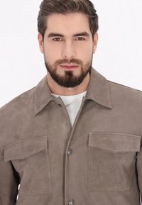 Braune Wildlederjacke mit Kragen, zwei Brusttaschen und Druckknopfverschluss. Weiche Textur, dezente Nähte und minimalistisches Design.