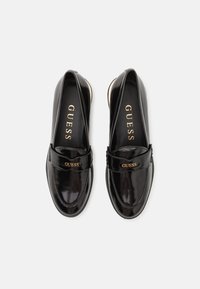 Mocasines de charol negro con un tacón bajo, que presentan una banda frontal y el logo dorado "GUESS". Textura suave y diseño de punta redondeada.