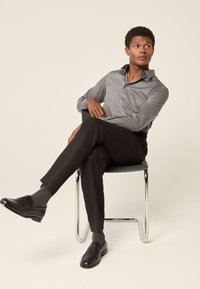 Szürke gombos ing, fekete szabott nadrág, fekete mokaszin és szürke zokni. A modell egy modern széken ül, laza pózban. Világos háttér.