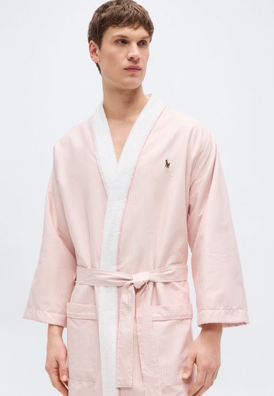 Νεαρός άνδρας που φοράει ρόμπα με ανοιχτό ροζ ρίγες, με λευκές λεπτομέρειες και δέσιμο στη μέση, κοιτάζοντας προς το πλάι με φόντο ένα απλό φόντο.