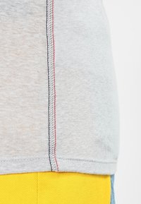 Gray cotton T-shirt with contrasting red and blue stitching along the side seam. Fabric has a soft texture. Yellow fabric layer visible below.