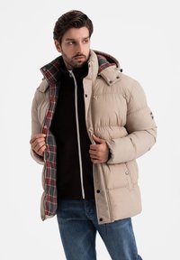 Beige puffjacka med mönstrad foder, dragkedjestängning och justerbar huva; har en slät yta och kviltad textur.