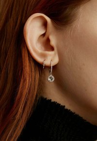 Pendientes de plata con un pequeño gema de tipo stud y un aro que presenta una piedra colgante más grande, sobre un fondo de tela negra texturizada.