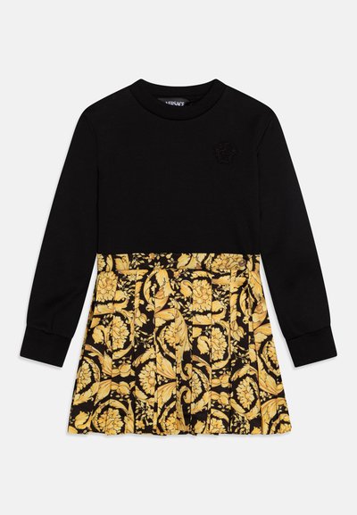 Juoda suknelė ilgomis rankovėmis su klostuota geltona gėlėta sijonu, su iškilmingu raštu ant krūtinės ir elastinėmis rankovių apykaklėmis.