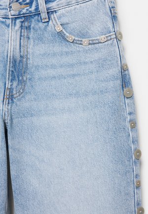 Svetlo modri jeansi iz jeansa z metalnimi gumbi vzdolž zunanje stranske šivne linije in majhnimi dekorativnimi zakovicami na robu sprednjega žepa.