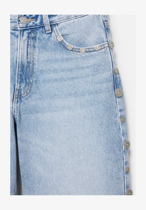 Svetlo modri jeansi iz jeansa z metalnimi gumbi vzdolž zunanje stranske šivne linije in majhnimi dekorativnimi zakovicami na robu sprednjega žepa.
