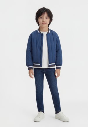 Ung person som står, iført en blå polstret jakke, hvit skjorte, blå jeans og hvite joggesko mot en ensfarget bakgrunn.