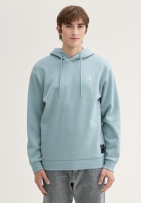 Felpa blu chiaro con tasca frontale, cordini regolabili e polsini a coste. Presenta un piccolo logo sul petto e un'etichetta in tessuto all'orlo.