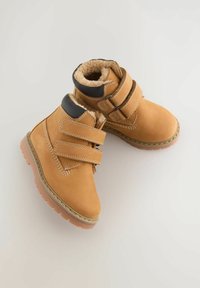 Braune Wildleder-Ankle Boots mit zwei Klettverschlüssen, warmem Kunstfellfutter, Gummisohle und dekorativen Nähdetails.