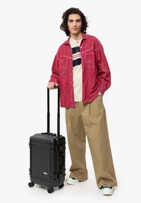 Valigia rigida nera con manico telescopico e quattro ruote, posizionata accanto a una persona che indossa una camicia rossa e pantaloni beige.