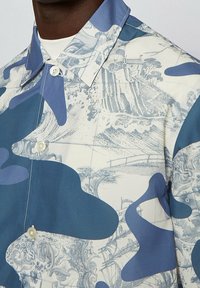 Lähikuva napitetusta sinivalkoisesta paidasta, jossa on abstrakteja ja yksityiskohtaisia maisemakuvioita.