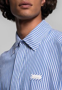 Camicia a righe blu e bianche con colletto button-down e tessuto testurizzato. Presenta un'etichetta bianca sul petto con il nome del marchio.