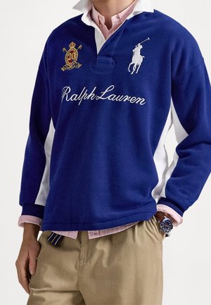Mężczyzna noszący niebieską koszulkę rugby Ralph Lauren na różowej koszuli z kołnierzykiem, beżowych spodniach i zegarek na rękę z niebieską tarczą.