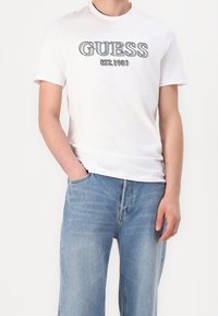 Weißes Baumwoll-T-Shirt mit dem Aufdruck "GUESS EST. 1981" in fetten, dunklen Buchstaben über der Brust, kombiniert mit hellblauen Denim-Jeans.