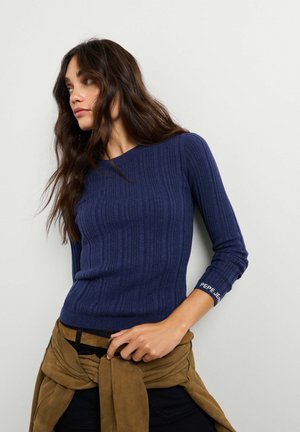 Giovane donna con capelli lunghi e mossi che indossa un maglione blu a coste aderente e una giacca marrone legata intorno alla vita, guarda di lato.