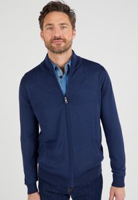 Marineblaues, zip-up Cardigan aus weichem Strickstoff mit hohem Kragen und geripptem Saum. Verfügt über Seitentaschen und ein aufgeknöpftes Jeanshemd darunter.