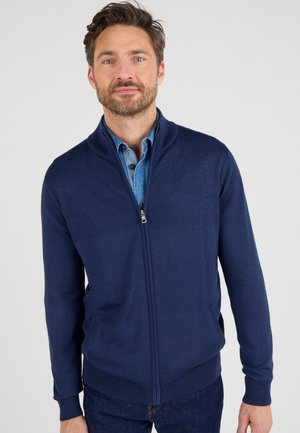 Cárdigan azul marino de cremallera en un suave tejido de punto con un alto cuello y dobladillo acanalado. Presenta bolsillos laterales y una camisa de mezclilla desabotonada debajo.