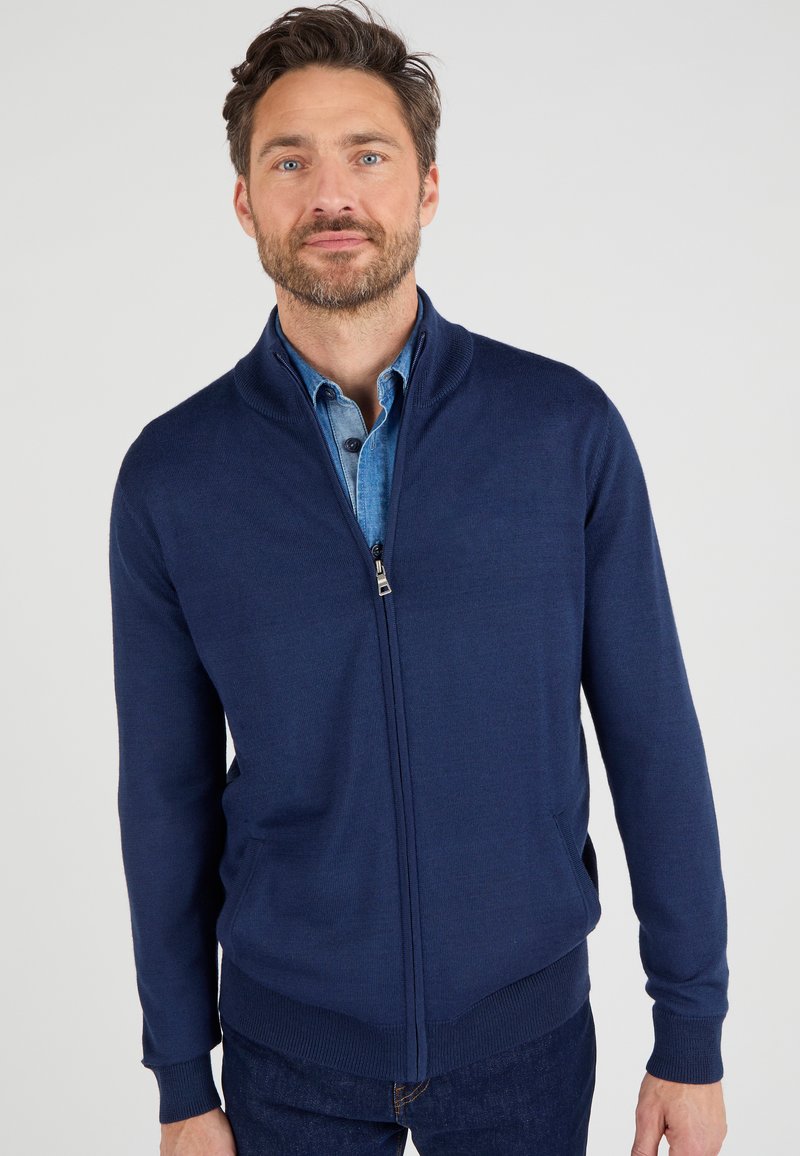 Marineblaues, zip-up Cardigan aus weichem Strickstoff mit hohem Kragen und geripptem Saum. Verfügt über Seitentaschen und ein aufgeknöpftes Jeanshemd darunter.