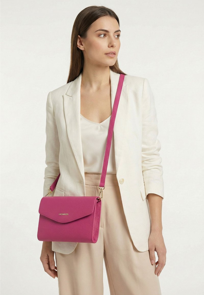 Mujer que lleva una chaqueta blanca y pantalones beige, lleva un bolso cruzado rosa Lazarotti con una correa larga sobre el hombro.