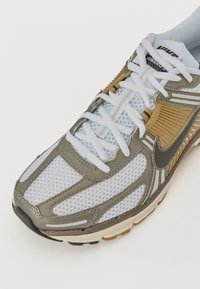 Sapatilha de corrida Nike com um cabedal em rede cinza claro, reforçado com detalhes em verde oliva e prateado, com atacadores brancos e uma sola acolchoada.