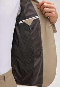 Innenansicht eines hellbraunen Blazers mit gemustertem schwarzem Futter, Innentasche mit Klappe und dem Etikett "Extra Slim Fit", getragen über einem weißen Hemd.