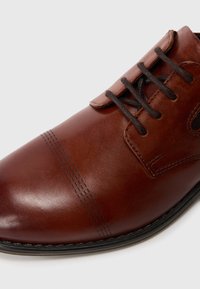 Chaussure en cuir marron à finition lisse, dotée de lacets foncés, d'un bout arrondi, de coutures décoratives et d'une semelle noire basse.