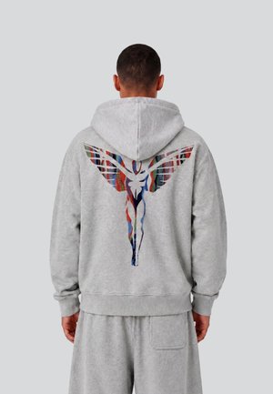 Mann trägt hellgrauen Hoodie und Shorts mit farbigem abstraktem Flügelmotiv auf dem Rücken, steht vor einfarbigem grauem Hintergrund.