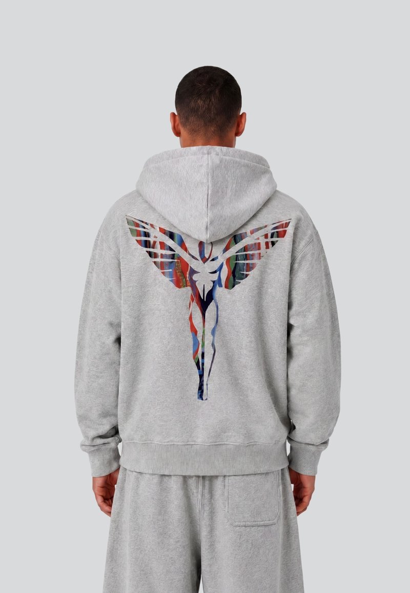 Homme portant un sweat à capuche gris clair et un short avec un motif abstrait coloré représentant une silhouette ailée dans le dos, debout devant un fond gris uni.