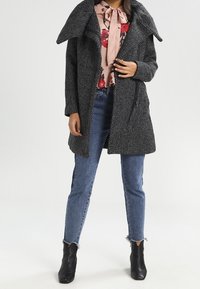 Grå tekstureret frakke med stor krave og åben front, parret med en blomsterbluse og blå jeans, båret med sorte ankelstøvler.