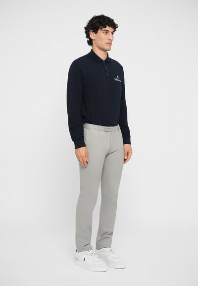 Camisa polo de manga larga en color navy con cuello y logo, combinada con pantalones gris claro y zapatillas blancas, sobre una figura masculina de pie contra un fondo blanco.