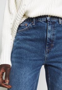 Ljusfärgad stickad tröja över mörkblå denimjeans. Jeans har en klassisk passform med synlig sömnad och fickor.