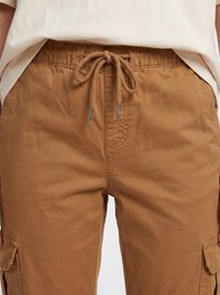 Parte centrale di una persona che indossa pantaloni cargo marroni con vita con coulisse annodata e una camicia beige infilata dentro.