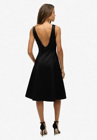 Schwarzes A-Linien-Kleid mit tiefem V-Ausschnitt hinten, aus glattem Material gefertigt. Verfügt über breite Träger und einen ausgestellten Rock, kombiniert mit schwarzen High Heels.