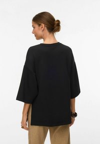 Ženska z lasmi v figi, oblečena v ohlapno črno majico s kratkimi rokavi in bež hlače, obrnjena stran, okrašena z uhanom, prstanom in zapestnico.