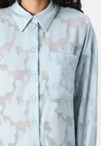 Camisa azul clara transparente com textura floral, gola, mangas longas e bolso frontal. Apresenta fecho de botão com botões azul claros.