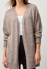 Długi, beżowy kardigan wykonany z miękkiej dzianiny. Posiada otwarty przód, ściągacze na mankietach i u dołu. Nosi go nad białą bluzką i czarnymi spodniami.