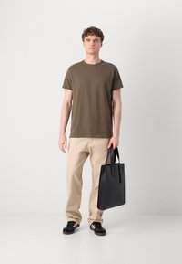Oliven grønn bomull T-skjorte, beige bukser, svart skinn totebag med korte håndtak, og svarte sneakers med hvite striper, stående foran en ensfarget bakgrunn.