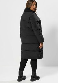 Czarna puchowa kurtka z wysokim kołnierzem, z teksturowanym futerkiem wewnątrz, bocznymi kieszeniami i długością do kolan. Noszona z czarnymi legginsami i botkami.
