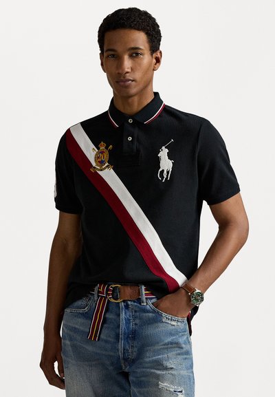 Polo shirt noir avec une rayure diagonale rouge et blanche, emblème 20 brodé et logo de cheval. Porté avec un jean bleu usé et une ceinture à motifs.