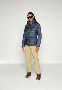 Γυναίκα με σκούρο μπλε καπιτονέ μπουφάν, μπεζ παντελόνι, καφέ μπότες και μαύρα γυαλιά ηλίου, που στέκεται σε μια απλή ανοιχτόχρωμη φόντο.