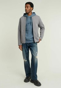 Giacca grigia sopra un felpa blu, jeans di denim con dettagli strappati e scarpe chunky nere. L'outfit presenta un design moderno e casual.