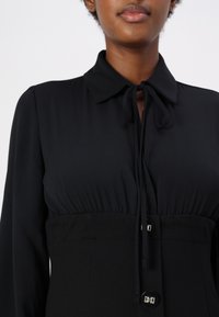 Blusa nera con colletto, dettaglio di fiocco davanti e bottoni decorativi. Presenta una vita aderente e una morbida texture del tessuto.