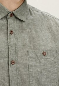 Camisa de lino gris con cuello abotonado, que presenta botones de plástico marrón y un bolsillo en el pecho con una tapa cuadrada. Textura sutil.