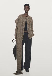 Cardigan in maglia marrone con una sciarpa lunga abbinata, caratterizzato da un design ampio, chiusura con bottoni e tessuto texturizzato; abbinato a pantaloni neri a gamba larga.