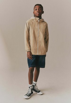 Noor poiss seisab, seljas beež kapuutsiga jakk, tumedad teksapüksid, valged sokid ja mustad kõrge säärega vabaajajalanõud, taust on ühevärviline.