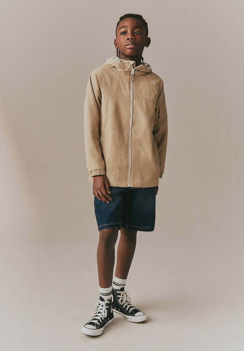 Junge steht und trägt eine beige Kapuzenjacke, dunkle Jeansshorts, weiße Socken und schwarze High-Top-Sneaker vor einem schlichten Hintergrund.